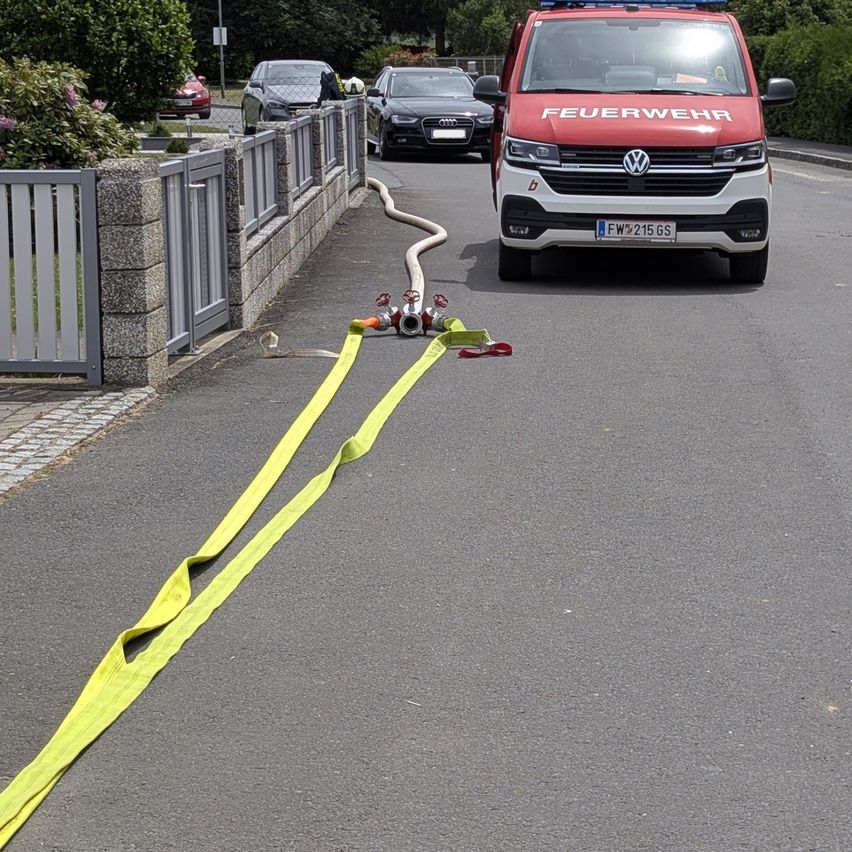 Ein roter Feuerwehrwagen steht am Straßenrand, mit einem Feuerwehrschlauch auf der Straße. Bäume und Pflanzen säumen die Straße. Eine Mauer befindet sich in der Nähe.