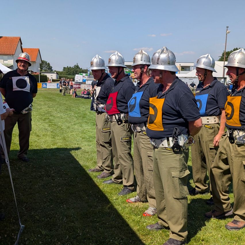 Eine Gruppe von Männern in Uniformen und Helmen steht auf einem Rasenfeld, möglicherweise bei einem Wettbewerb. Gebäude und Bäume sind im Hintergrund.