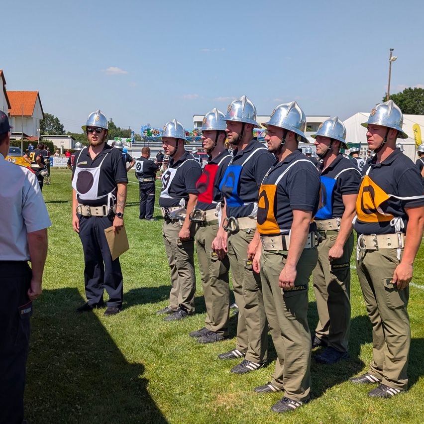 Eine Reihe von Männern in Uniform, jeder mit einem Helm und einer nummerierten Schürze, steht auf einem Rasenfeld. Ein Mann hält ein Klemmbrett. Im Hintergrund sind ein Haus und ein Auto zu sehen.