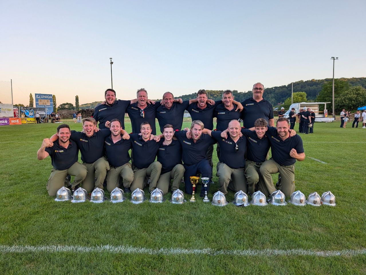 Eine Gruppe von Männern in schwarzen Shirts und khakifarbenen Hosen posiert für ein Foto auf einem Rasenfeld mit Trophäen vor ihnen.