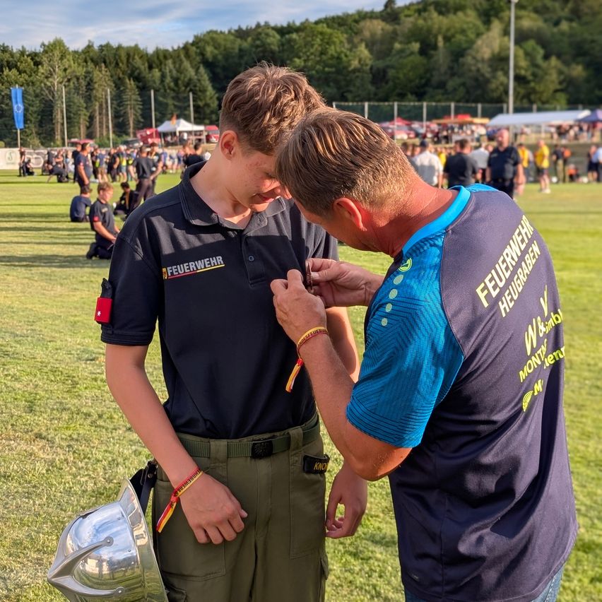 Zwei Männer stehen auf einem Grasfeld, einer in Uniform und der andere in einem T-Shirt, möglicherweise bei einer Feuerwehrveranstaltung. Im Hintergrund sitzen mehrere Personen, und mehr stehen in der Ferne. Bäume und eine blaue Fahne sind in der Ferne zu sehen.