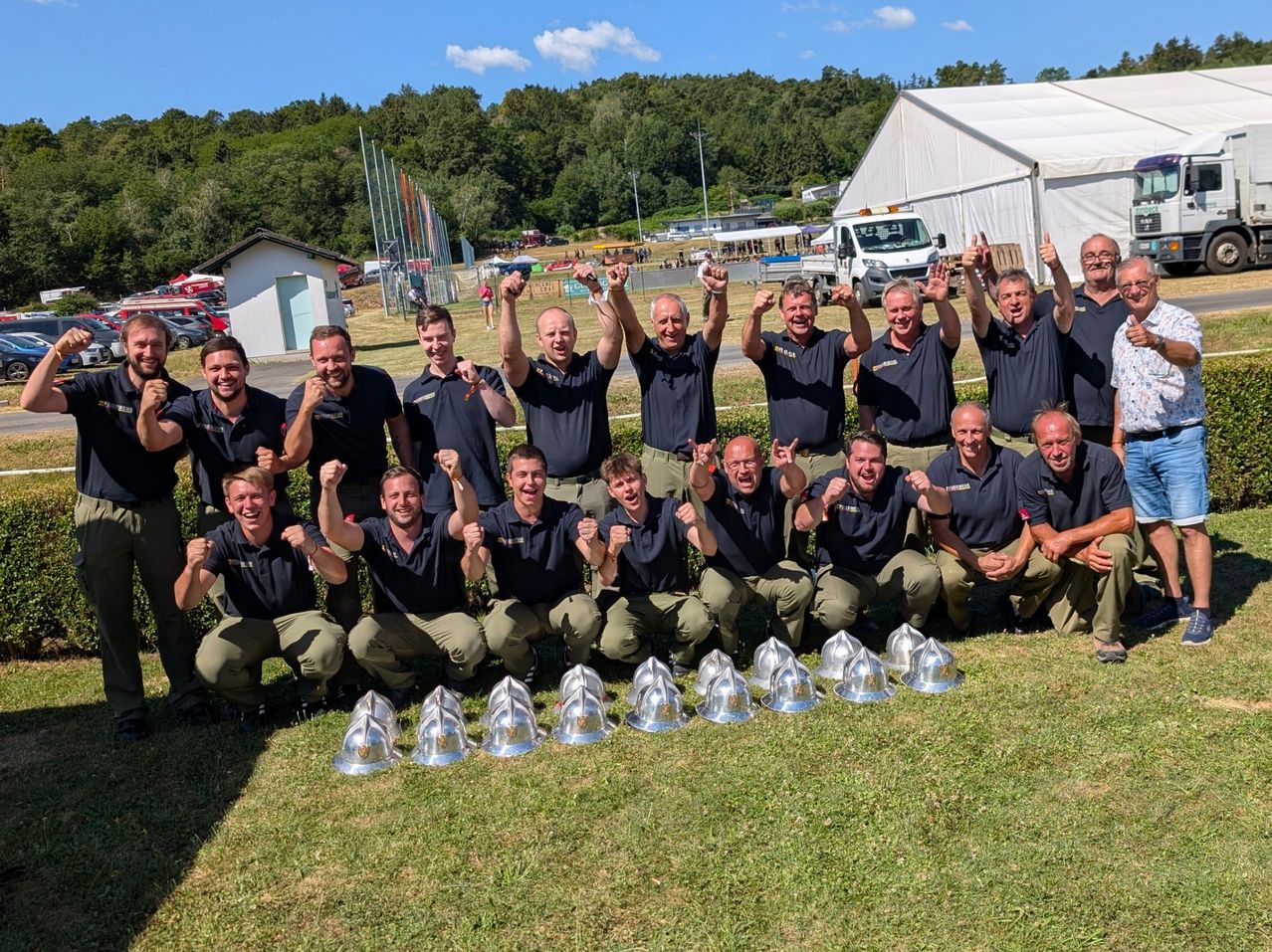 Eine Gruppe von Feuerwehrleuten posiert mit Helmen vor einem Zelt und Grünpflanzen. Sie tragen alle Uniformen und lächeln.