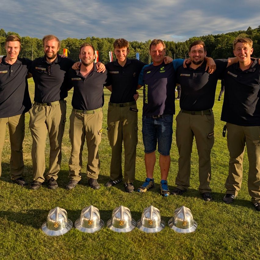 Sieben Männer in Uniform posieren für ein Foto auf einem Rasenfeld, mit Helmen davor. Sie lächeln für die Kamera unter einem bewölkten Himmel mit Bäumen im Hintergrund.