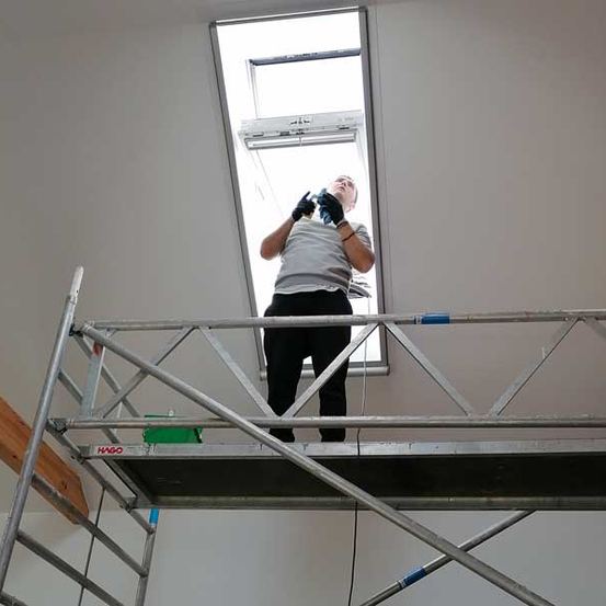 Ein Mann steht auf einem Gerüst und schaut durch ein offenes Dachfenster. Er trägt ein weißes Hemd, schwarze Hose und schwarze Handschuhe.