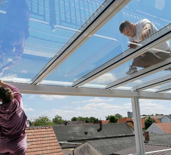 Zwei Personen arbeiten an einem Glasdach. Eine Frau reinigt das Glas und ein Mann repariert etwas. Sie stehen auf einem Dach mit Blick auf die Stadt unten.