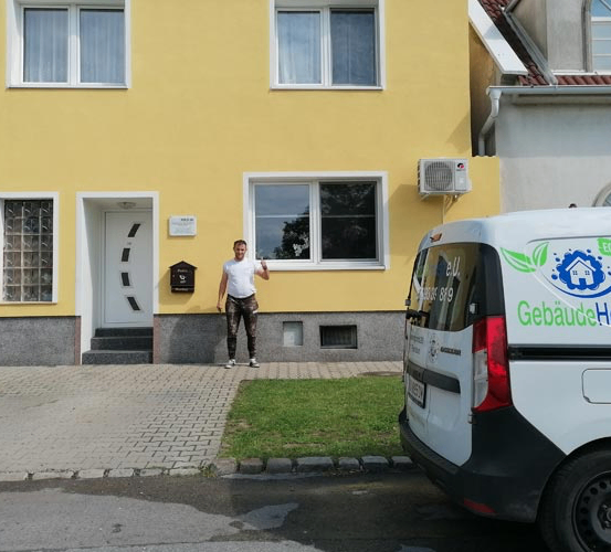 Ein Mann in Camouflage-Hosen winkt vor einem gelben Haus mit einem weißen Van davor.