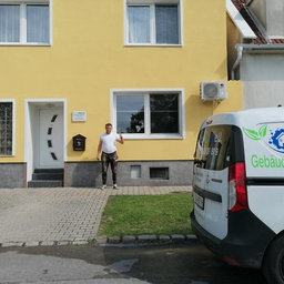 Ein Mann in Camouflage-Hosen winkt vor einem gelben Haus mit einem weißen Van davor.