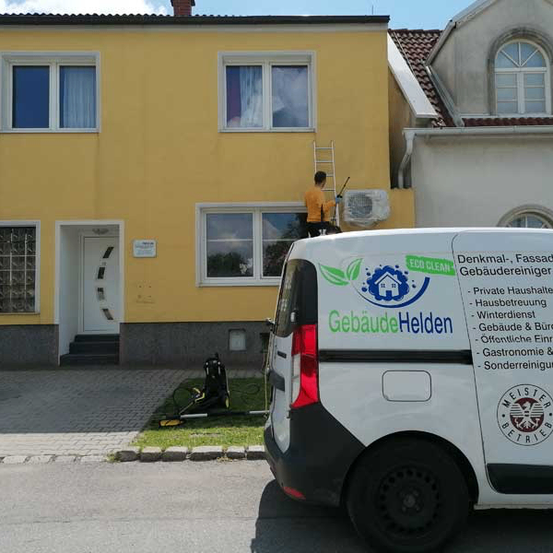 Ein Mann reinigt ein Fenster eines gelben Hauses. Ein weißer Van mit dem Text 'Gebaude Helden' steht davor.