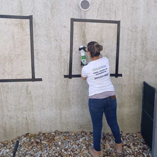 Eine Frau in einem weißen T-Shirt reinigt einen Fensterrahmen an einer Betonwand mit einer Sprühflasche. Sie trägt Handschuhe und blaue Jeans. Die Wand hat einen schwarzen Rand.