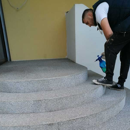 Ein Mann in einer Weste und Sneakers reinigt eine graue Betontreppe mit einer blauen Sprühflasche.