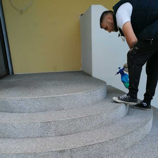 Ein Mann in einer Weste und Sneakers reinigt eine graue Betontreppe mit einer blauen Sprühflasche.