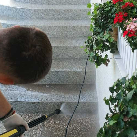 Ein Mann reinigt die Treppe mit einem Wasserschlauch, trägt ein weißes T-Shirt und Handschuhe, in der Nähe von Pflanzen mit roten Blumen.