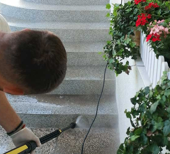 Ein Mann reinigt die Treppe mit einem Wasserschlauch, trägt ein weißes T-Shirt und Handschuhe, in der Nähe von Pflanzen mit roten Blumen.