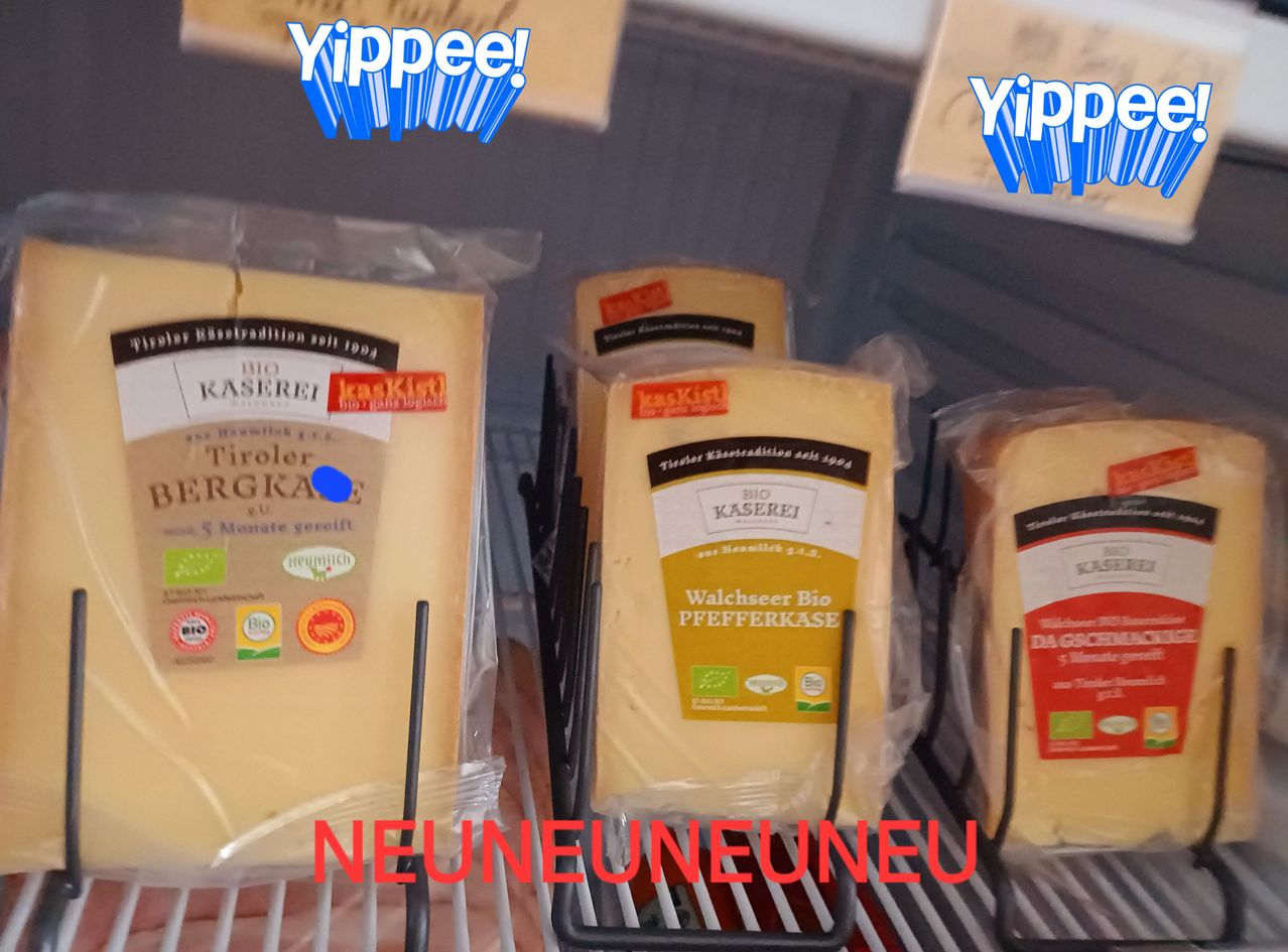 Mehrere Packungen Käse sind auf einem Regal ausgestellt. Der Käse ist in Plastikverpackungen mit Etiketten, die darauf hinweisen, dass er biologisch und von Tiroler Bergkäse und Walchseer Bio Pfefferkase stammt. Das Bild hat eine verspielte Überlagerung mit dem Text 'Yippee!' und 'NEUNEUNEUNEU'.