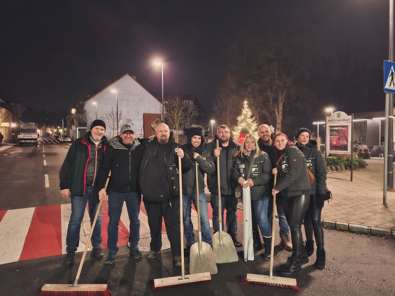 A group of people in winter attire are posing with their shovels in a snowy area at night. A Christmas tree and houses are visible in the background.