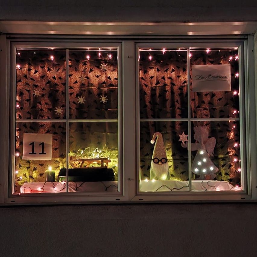 Ein Fensterdisplay mit Schneeflocken und Lichtern, einer Kerze und einem Schild mit der Nummer 11. Ein Gnom und Engelfiguren sind im Fenster.