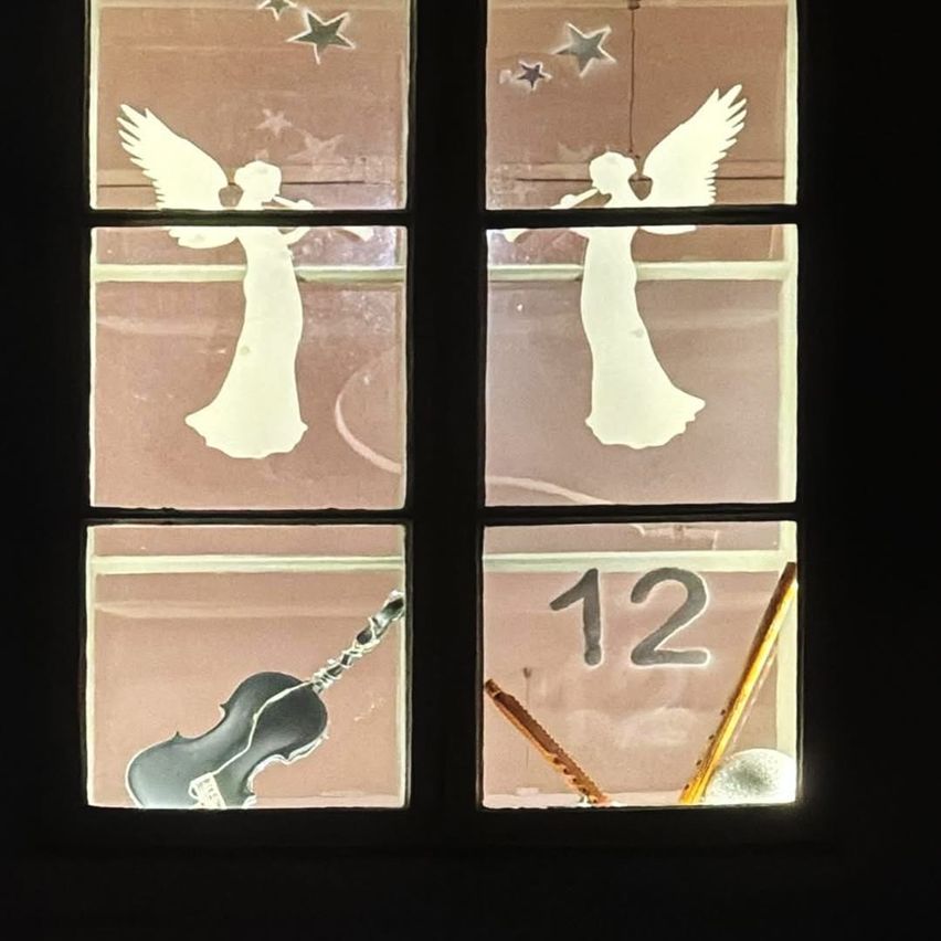 Zwei Engel, die Instrumente spielen, sind auf einem Fenster zu sehen. Der Engel auf der linken Seite spielt eine Flöte, während der Engel auf der rechten Seite eine Trompete spielt. Unten ist eine Violine zu sehen, und die Nummer 12 ist sichtbar.