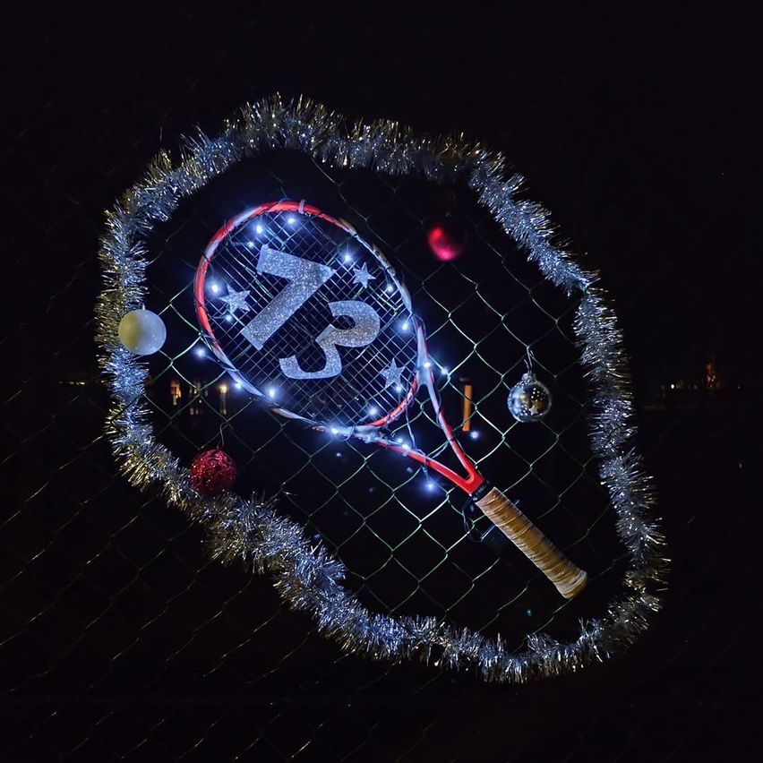 Ein Tennisschläger ist mit Lichtern und Ornamenten geschmückt. Es ist von einer Tinselgirlande in Herzform umgeben. Der Schläger steht auf einem Zaun.