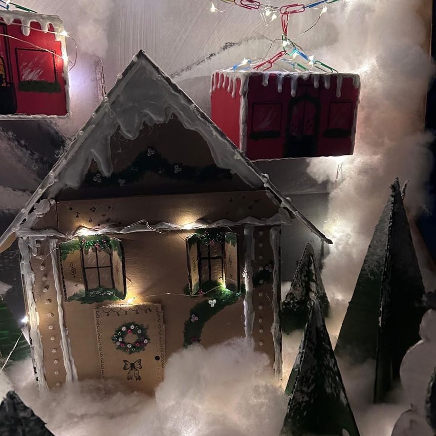 Ein Lebkuchenhaus mit schneebedecktem Dach und Fenstern, die mit Weihnachtskränzen und Lichtern geschmückt sind. Es ist von künstlichem Schnee und Bäumen umgeben, mit anderen festlich geschmückten Häusern im Hintergrund.