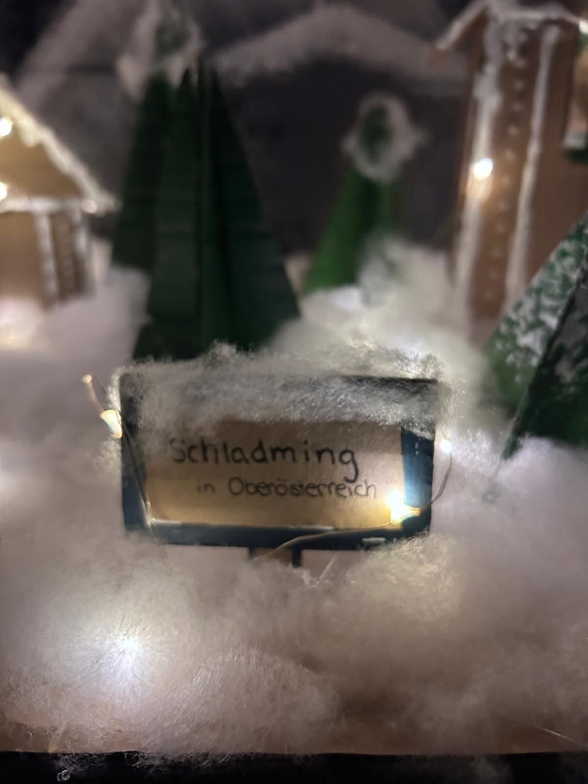 Eine Winterszene mit einem Schild, das 'Schladming in Oberosterreisch' besagt. Das Gebiet ist schneebedeckt mit Miniatur-Weihnachtsbäumen und Häusern im Hintergrund.