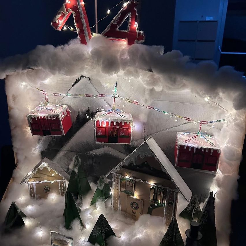 Eine Weihnachtsszene mit einer Gondelbahn über einer verschneiten Landschaft. Die Gondeln sind rot mit Weihnachtsdekoration. Unten sind schneebedeckte Bäume und Häuser mit Lichtern. Die Nummer 14 ist oben in roten Lichtern.