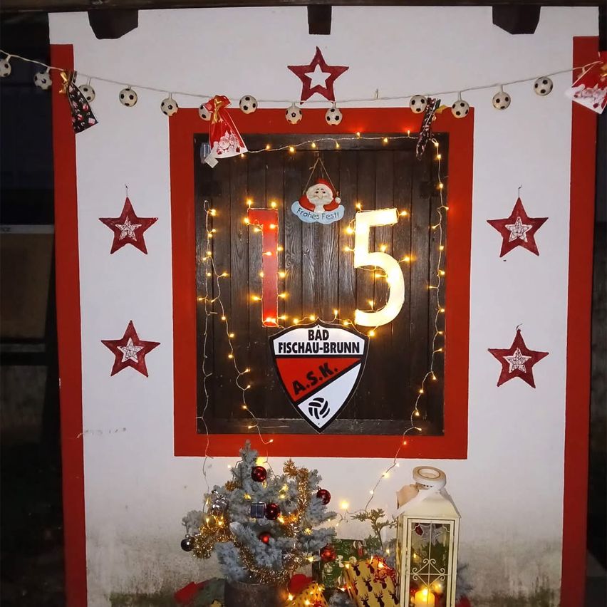 Eine dekorierte Holztür mit der Nummer 15, dem Bad Fischau-Brunn-Emblem und festlichen Lichtern. Ein Weihnachtsbaum, eine Laterne und Fußballbälle davor.