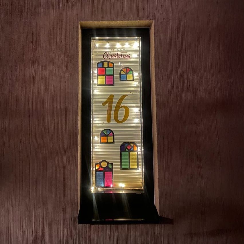 Ein gerahmtes dekoratives Stück mit der Nummer 16 und der Aufschrift Weihnachten. Es zeigt eine Reihe kleiner Fenster mit mehrfarbigen Glasscheiben, die von Lichtern beleuchtet werden.