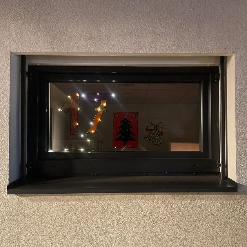 Ein schwarzer Fensterrahmen mit einem Glasfenster reflektiert Weihnachtsdekorationen. Eine beleuchtete Nummer 17 und andere Weihnachtsornamente sind auf der Scheibe zu sehen.