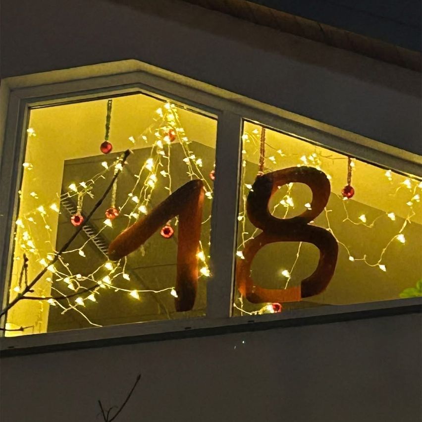 Ein Fenster mit zwei großen Holzzahlen, 1 und 8, umgeben von Weihnachtslichtern und Ornamenten. Das Licht spiegelt sich im Glas.