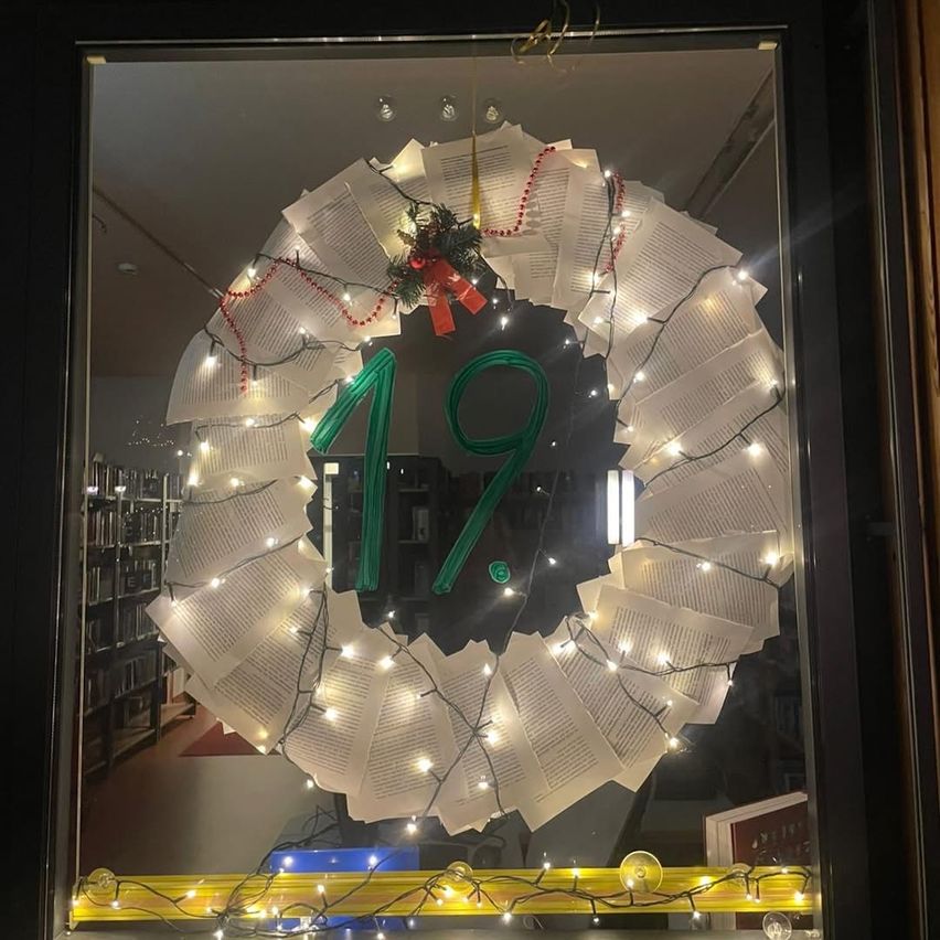 Ein Kranz aus Buchseiten mit Weihnachtslichtern in einem Bibliotheksfenster, mit der Nummer 19 prominent angezeigt.