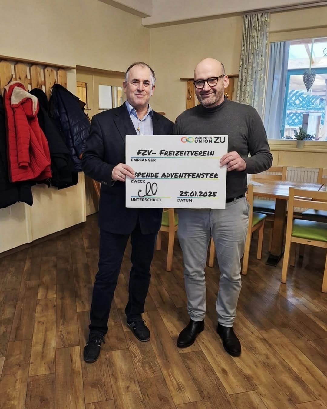 Zwei Männer in Anzügen halten einen Scheck und lächeln für die Kamera. Ein Jacke hängt hinter ihnen, und ein Tisch mit Stühlen ist im Hintergrund.