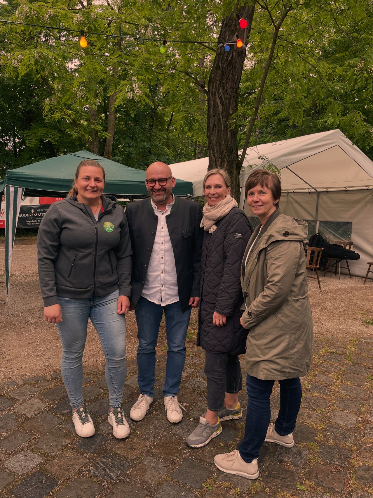 Four people are standing together outdoors in a park. They are all smiling and appear to be posing for a photograph. Behind them, there are tents set up.