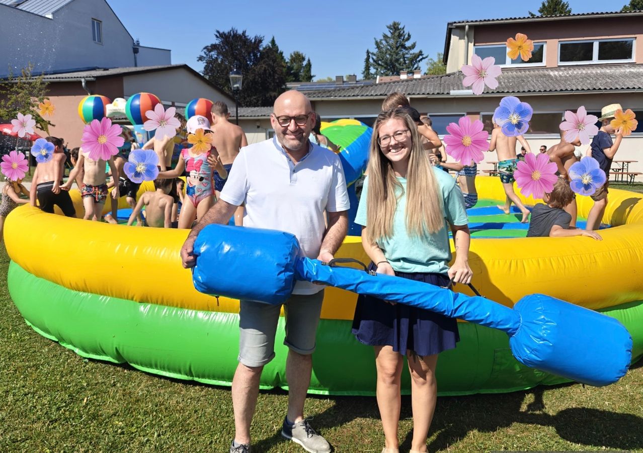 Ein Mann und eine Frau stehen vor einem großen aufblasbaren Hüpfburg mit einer blauen Tasche. Dahinter sind Menschen, die die wassergefüllte Hüpfburg genießen.