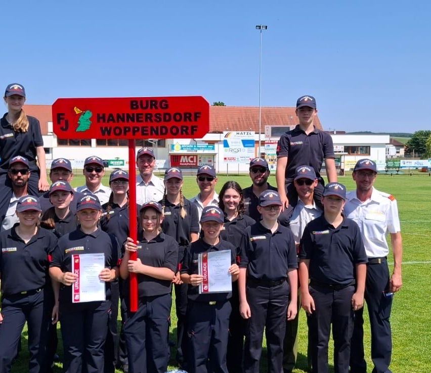 Eine Gruppe von Menschen in Uniform posiert für ein Foto im Freien. Ein Schild steht Burg Hannersdorf Woppendorf.