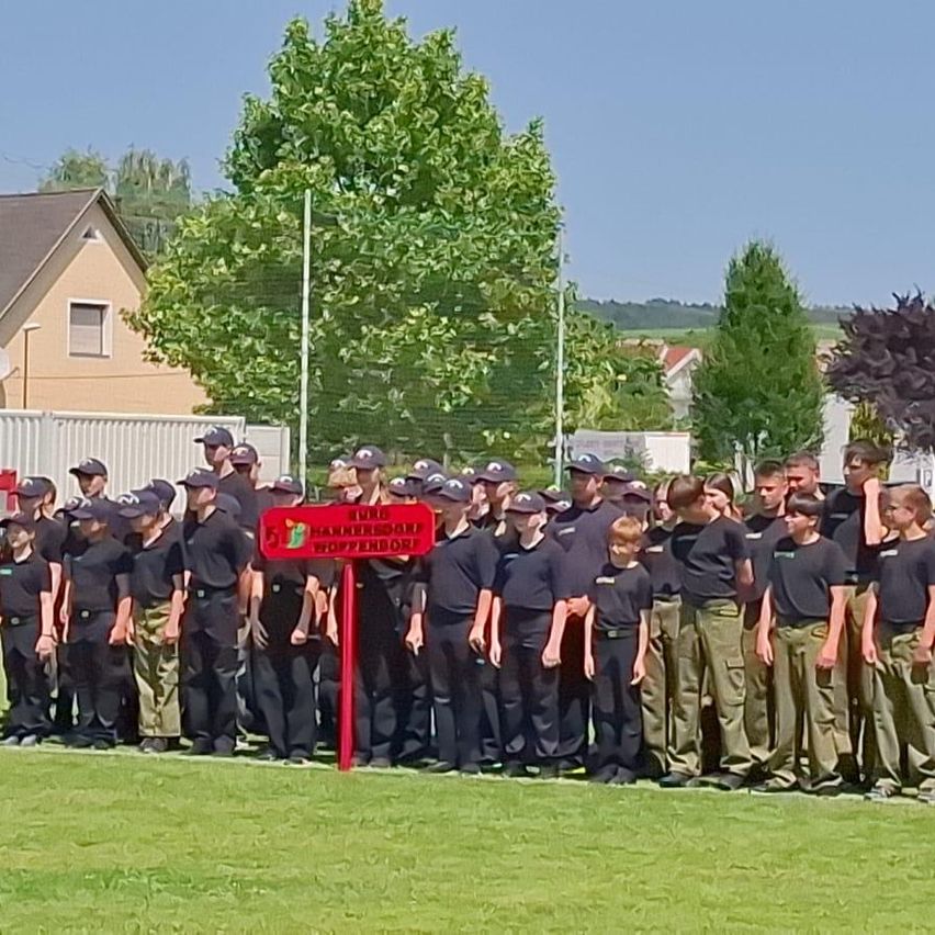 Eine Gruppe uniformierter Personen steht auf einem Feld mit einem Schild, das 'Würz Wipperdorf' besagt. Sie stehen in einer Reihe, mit Häusern und Bäumen im Hintergrund.