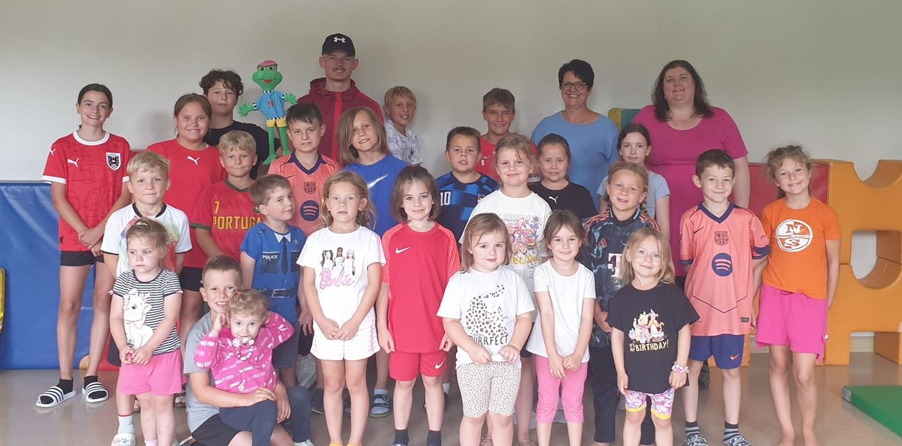 Eine Gruppe von Kindern und Erwachsenen posiert für ein Foto. Die Kinder tragen verschiedene farbige T-Shirts, während Erwachsene hinter ihnen stehen. Der Raum ist weiß gestrichen.