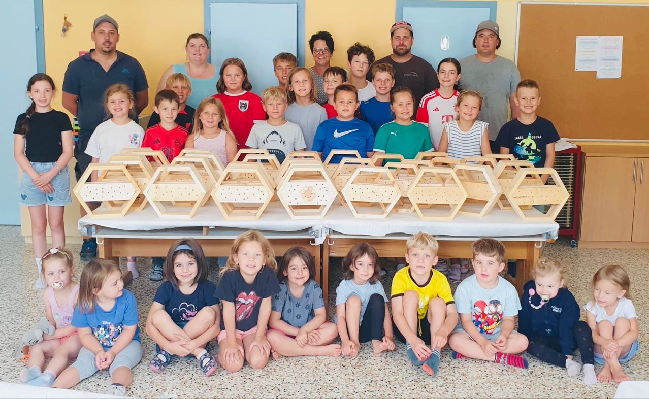 Eine Gruppe von Kindern und Erwachsenen versammelt sich lächelnd um einen Tisch mit sechseckigen Holzkisten. Einige tragen Sporttrikots und einige tragen Brillen. Der Raum hat gelbe Wände und blaue Türen.