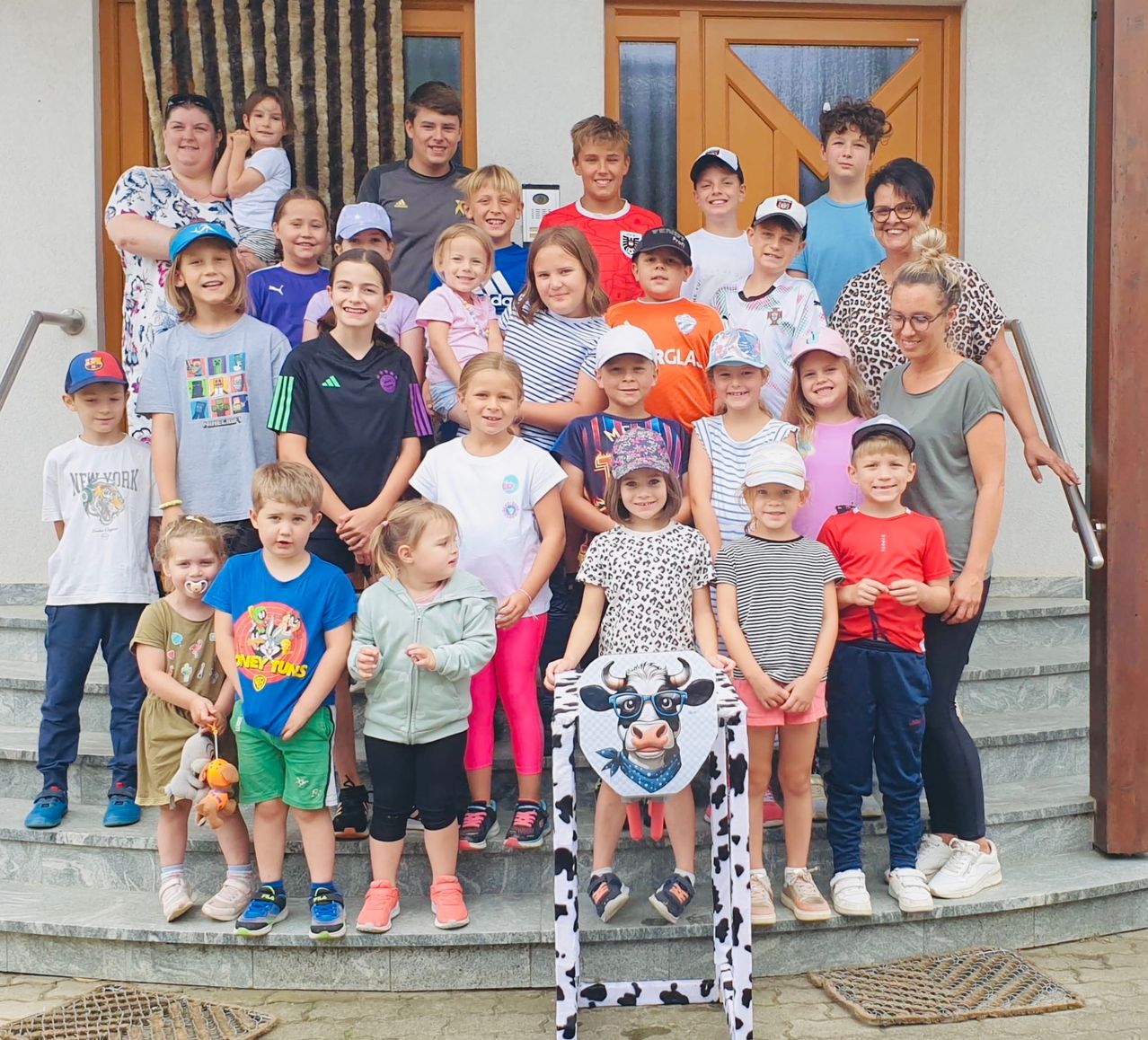 Eine Gruppe von Kindern und Erwachsenen steht auf den Stufen eines Gebäudes. Einige tragen Mützen und Turnschuhe. Ein Mädchen hält einen Kuhausschnitt vor sich. Hinter ihnen hält eine Frau ein Baby auf der Schulter.