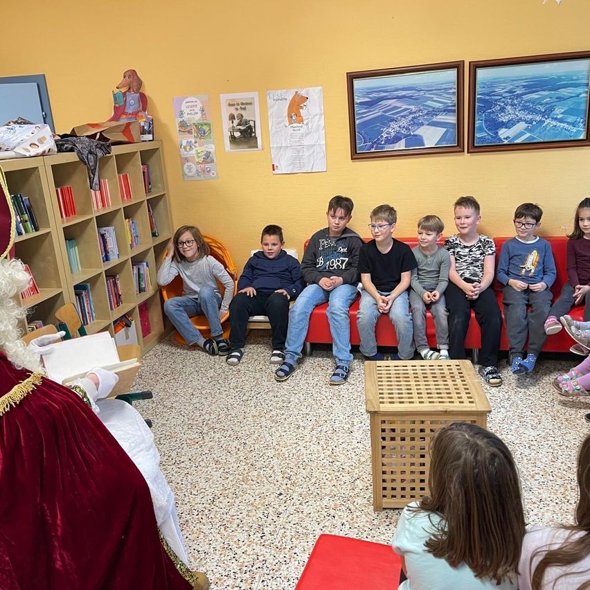 Eine Gruppe von Kindern hört einer Geschichte zu, die von einer als Weihnachtsmann verkleideten Person in einem Raum mit gelben Wänden und einem mit Büchern gefüllten Bücherregal vorgelesen wird.