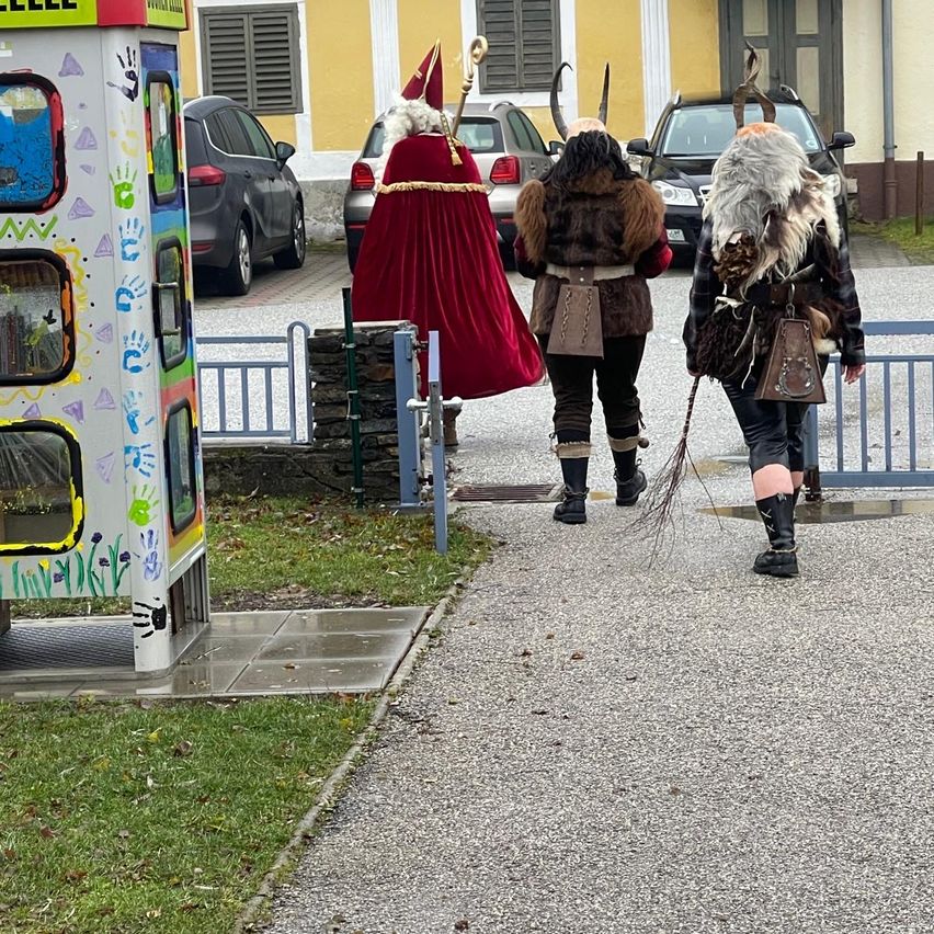 Drei Menschen in Wikingerkostümen gehen auf einem Stadtplatz, an dem ein farbenfroher Kiosk und geparkte Autos im Hintergrund stehen.