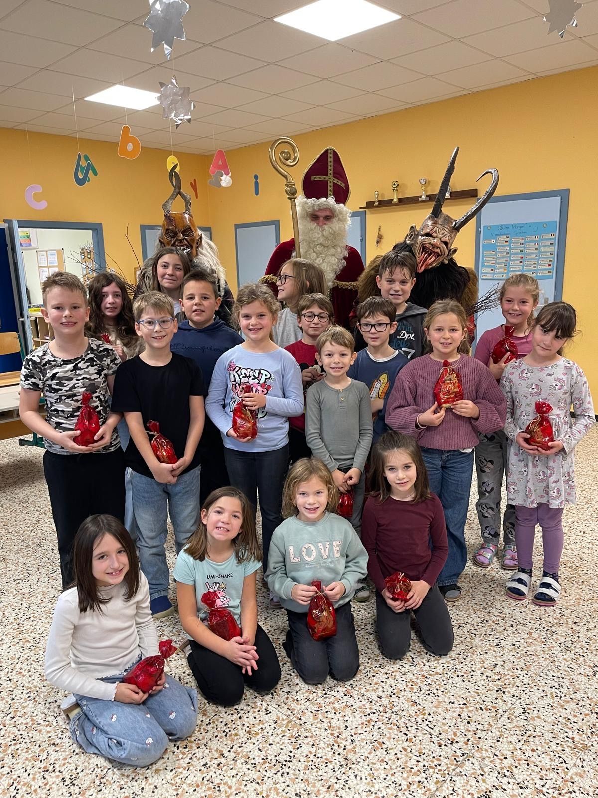 Eine Gruppe von Kindern mit roten Tüten posiert für ein Foto mit zwei Erwachsenen, die als Weihnachtsmann und Krampus verkleidet sind, in einem Raum mit gelben Wänden und einem gemusterten Boden.