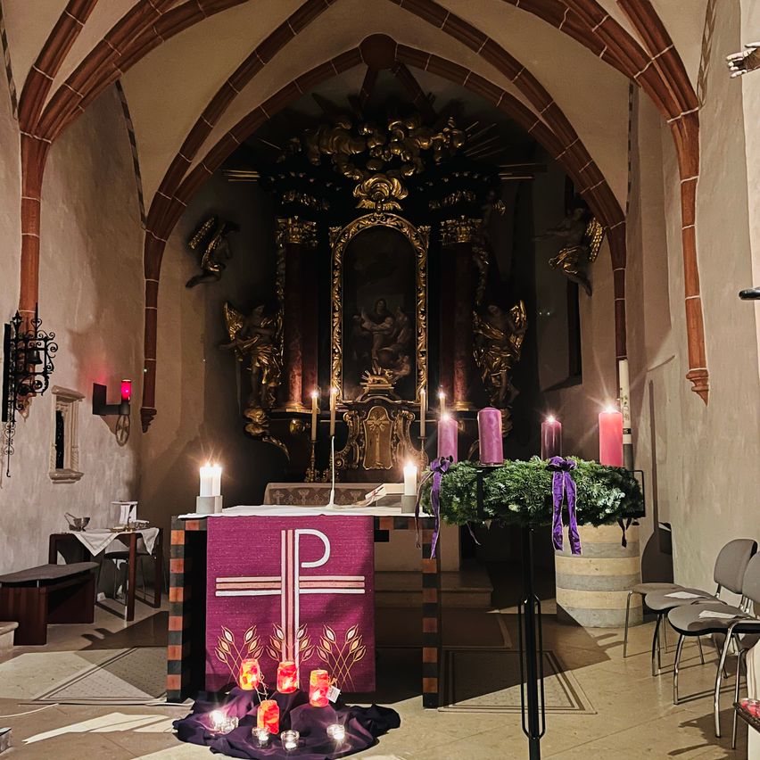 Eine schwach beleuchtete Kapelle mit einem Kreuz und Kerzen auf dem Altar. Der Altar hat einen violetten Stoff mit einem Kreuz und Getreidesymbolen. Es gibt Stühle und einen Holztisch.