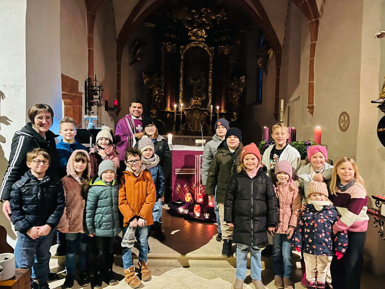 Eine Gruppe von Kindern und Erwachsenen steht vor einem Altar mit Kerzen in einer Kirche.