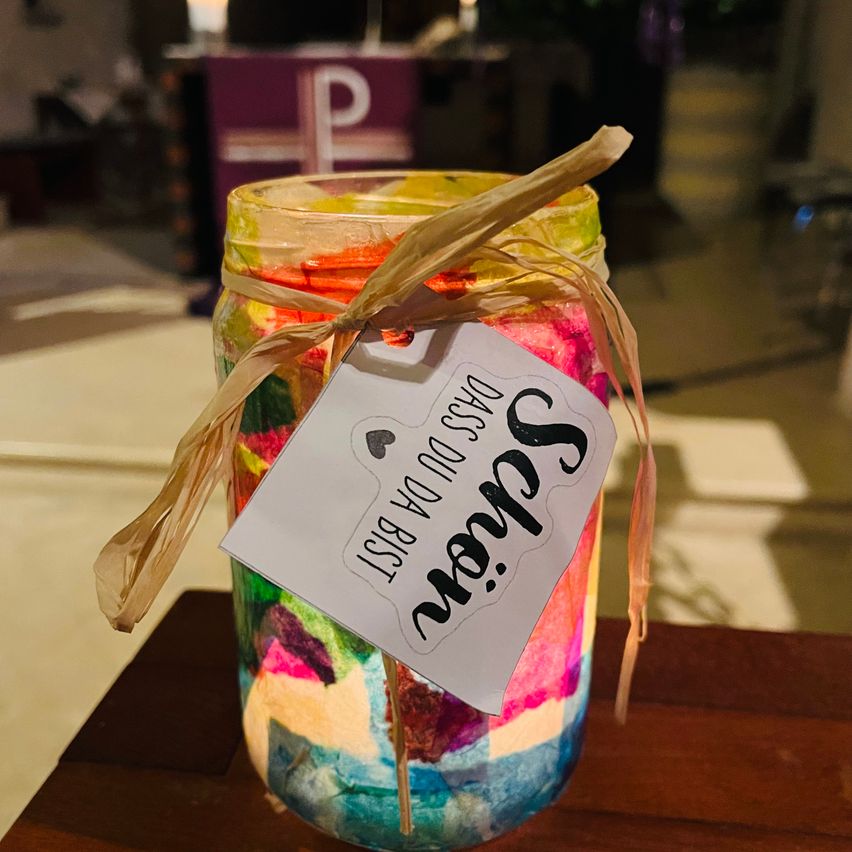 Ein farbenfrohes Glas mit einem Etikett, auf dem 'Funkeln' steht, ist mit mehrfarbigem Papier gefüllt. Mit einem Band befestigt, steht es auf einem Holztisch.