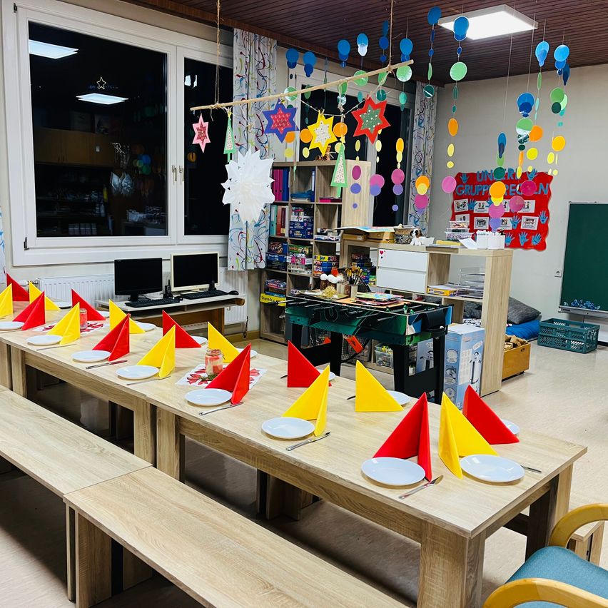 Ein Klassenzimmer mit langen Holztischen, die mit Tellern, bunten Servietten und Besteck gedeckt sind. Der Raum hat eine bunte Hängedekoration, eine Tafel und einen Heizkörper.