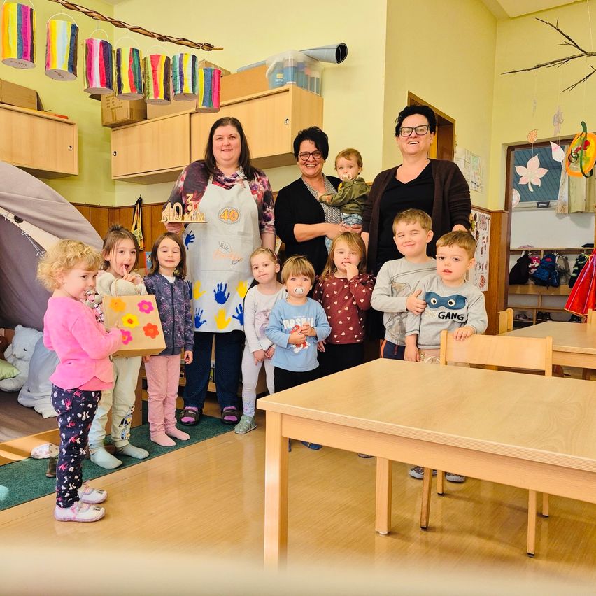 Eine Gruppe von Erwachsenen und Kindern in einem Raum, einige halten Geschenke und lächeln für ein Foto. Eine Frau in einer weißen Schürze hält einen Kuchen mit der Nummer 40.