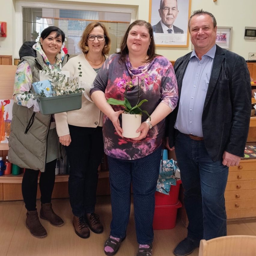 Vier Erwachsene stehen drinnen in der Nähe eines Fensters und lächeln für ein Foto. Zwei Frauen halten Pflanzen; eine hat Orchideen und die andere Grünpflanzen. Ein Mann und eine Frau stehen in der Mitte, wobei die Frau eine Topfpflanze hält. Ein gerahmtes Bild eines Mannes ist an der Wand angebracht.