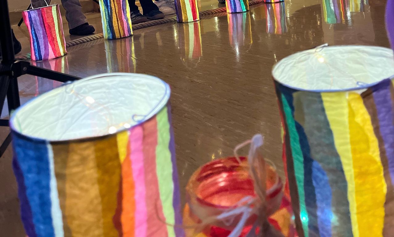 Nahaufnahme von bunten Papierlaternen auf einem Holzboden, Reflexionen von Füßen, einem Schuh und einem Armband.