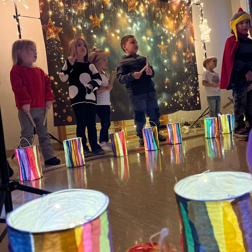 Kinder stehen auf einer Bühne und halten Trommeln und bunte Eimer. Ein Hintergrund zeigt Sterne und Lichter. Ein weiteres Kind steht an der Seite.