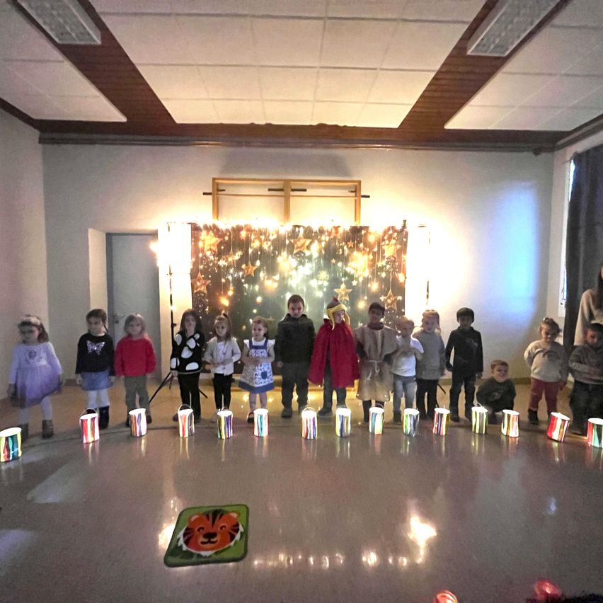 Eine Gruppe von Kindern steht im Kreis und hält jeweils eine brennende Kerze. Sie befinden sich in einem Raum mit weißer Decke und dekorativem Hintergrund. Erwachsene stehen in der Nähe, und auf dem Boden liegt ein Löwenteppich.